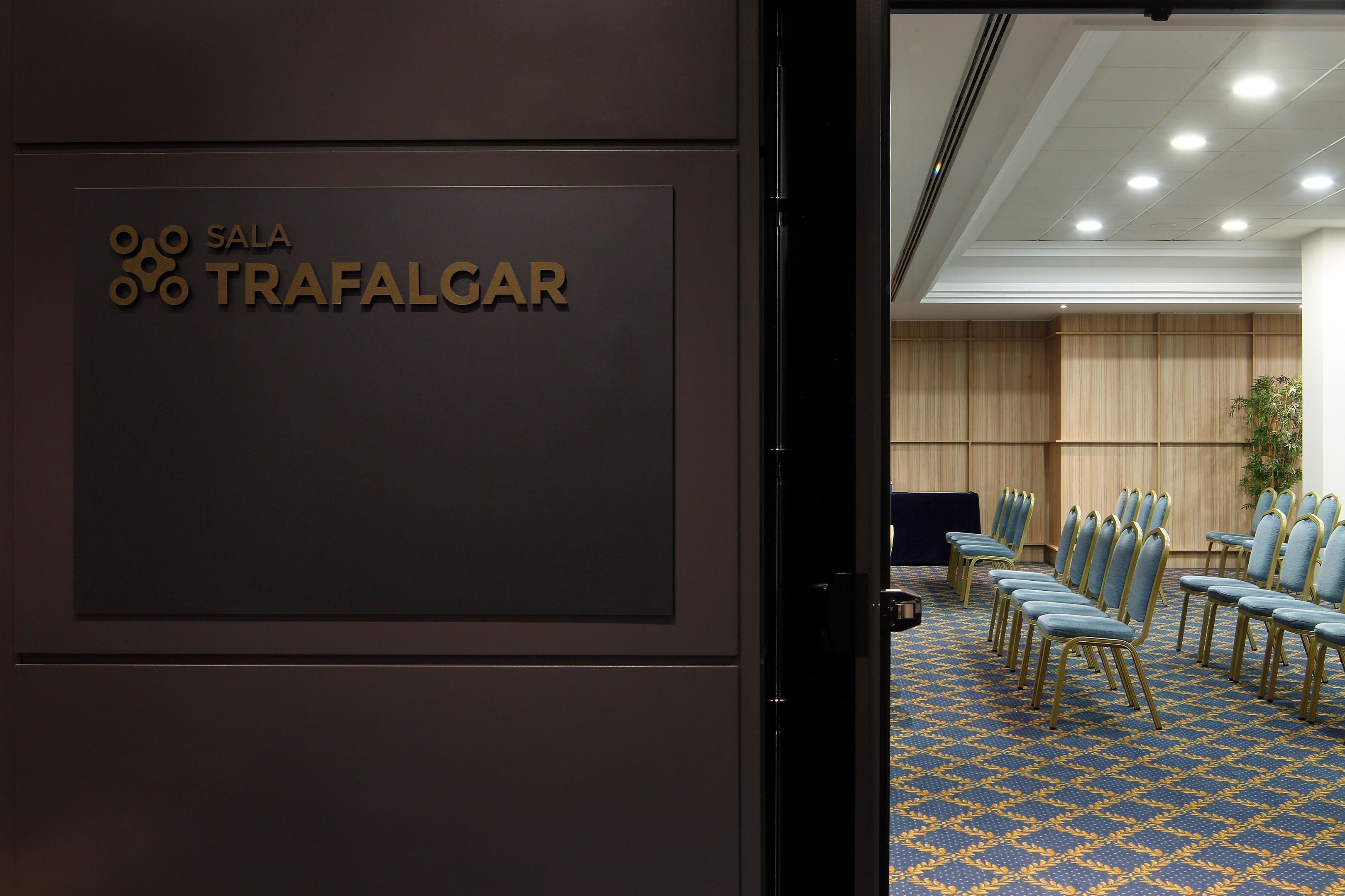 Una sala conferenze con un cartello che indica “SALA TRAFALGAR” e file di sedie blu con cornice dorata.