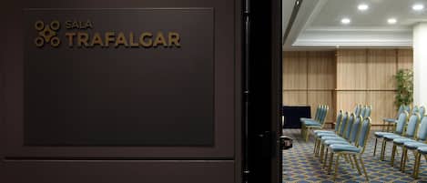 A conference room with a sign reading "SALA TRAFALGAR" and rows of blue chairs with gold frames.