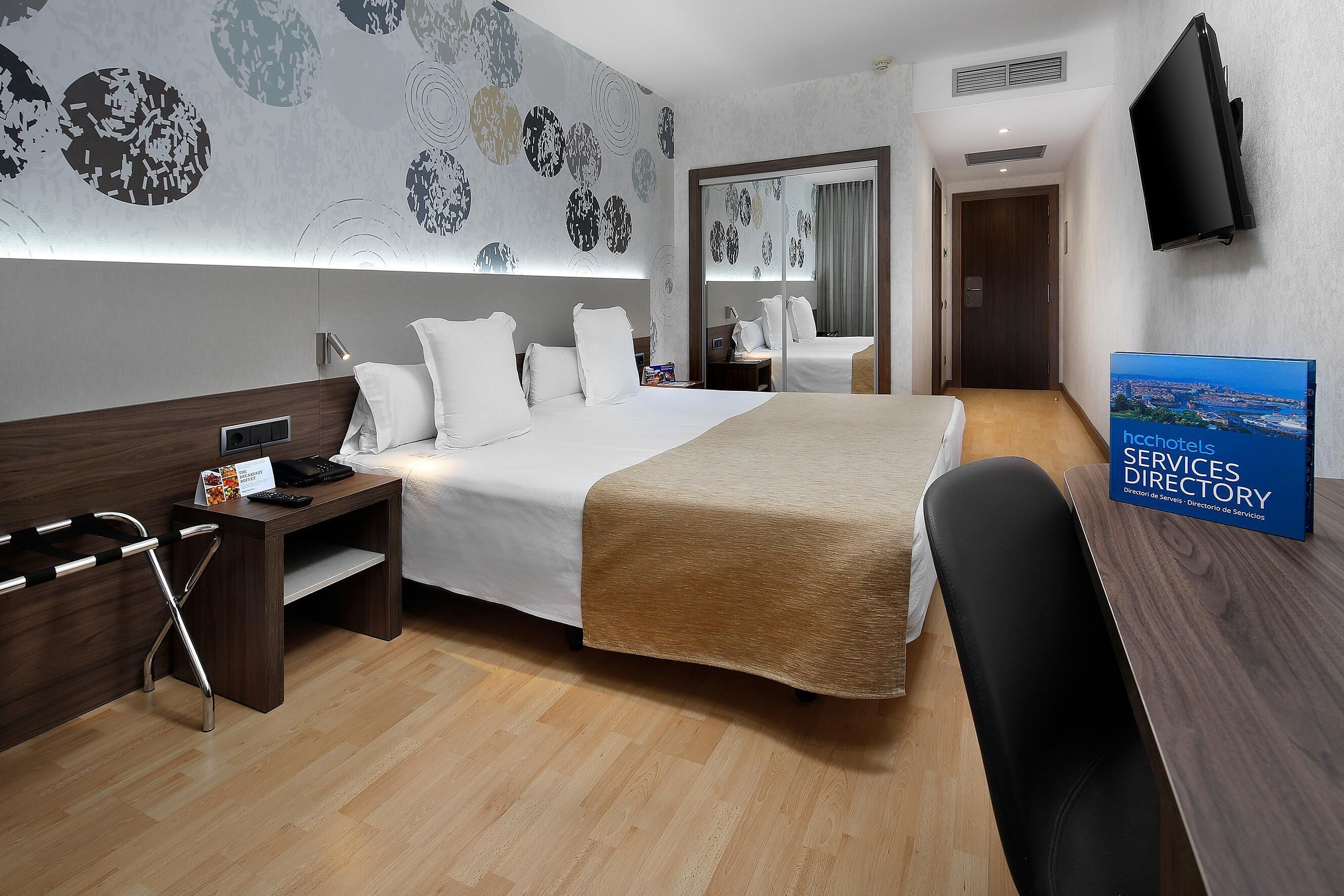 A hotel room with a large bed, bedside tables, a desk, a chair, a flat-screen TV, and a signboard.