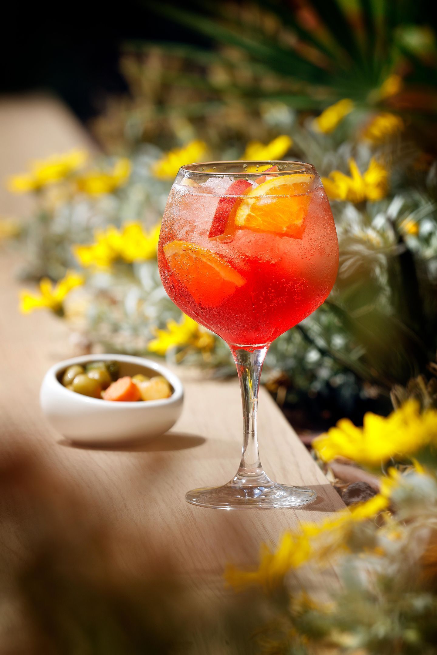 A cocktail glass with fruit slices on a wooden table, surrounded by yellow flowers and a small bowl of olives.