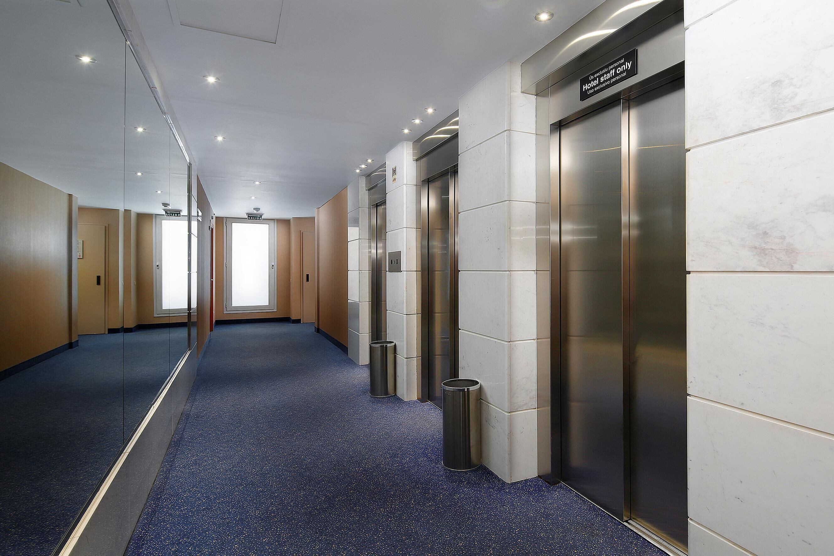 A hotel corridor with a blue carpet, mirrored walls, and two elevators.