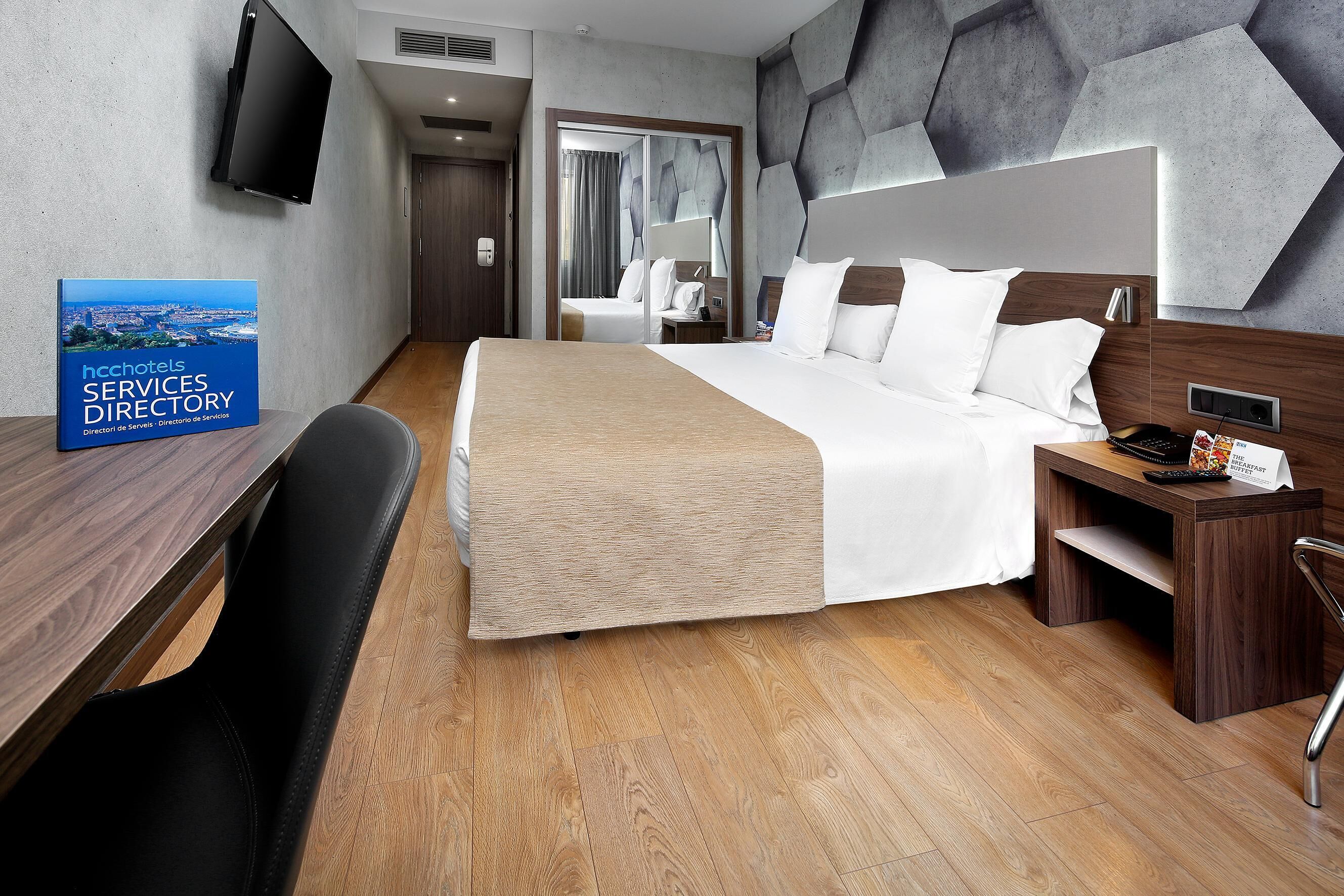 A hotel room with a large bed, a desk with a chair, a TV mounted on the wall, and a book titled "HCHOTELS SERVICES DIRECTORY" on the desk.