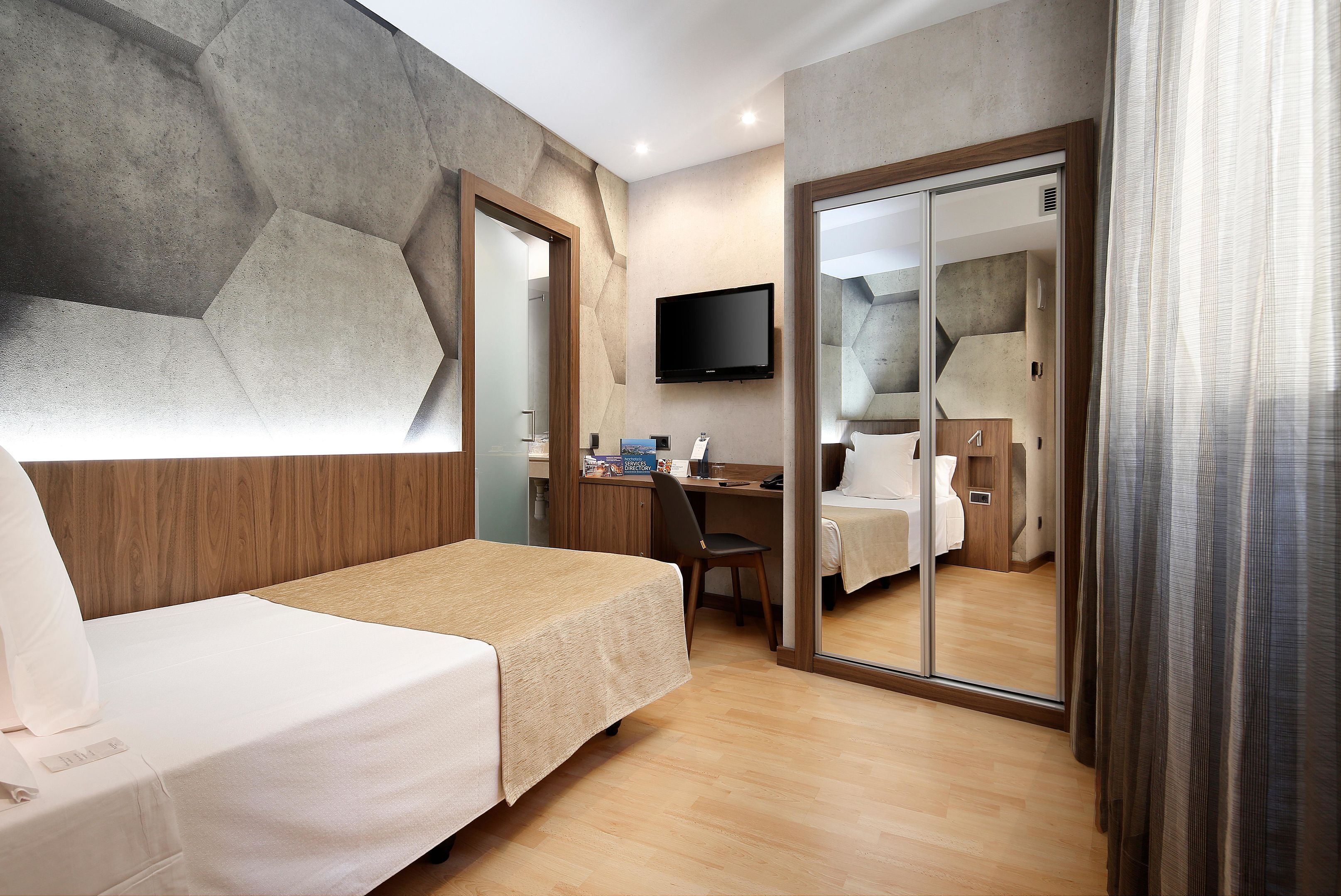 Quarto de hotel com cama, escrivaninha, cadeira, televisão e espelho.