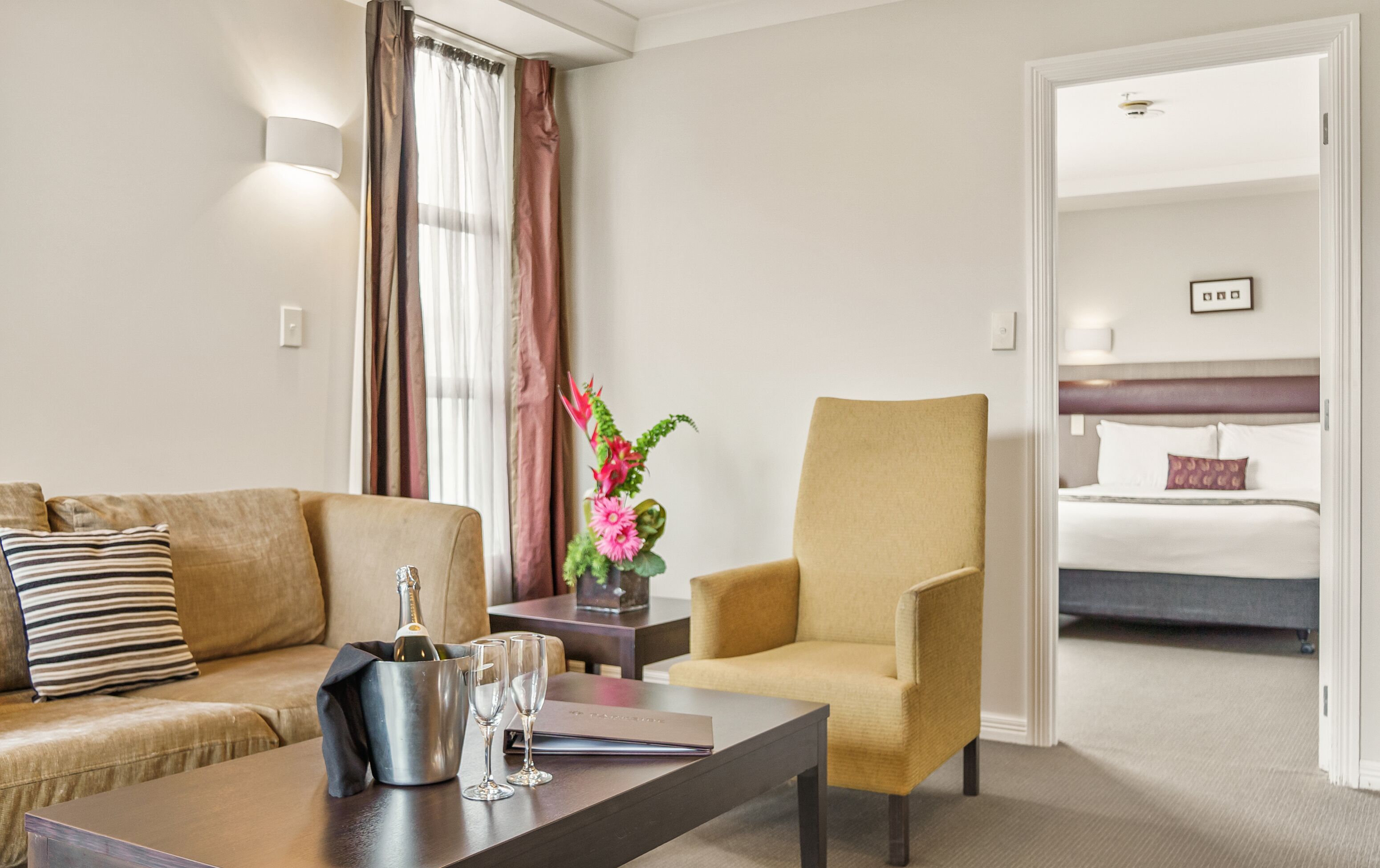 A hotel room with a sofa, armchair, a small table with a bottle and glasses, and a bed with a headboard.