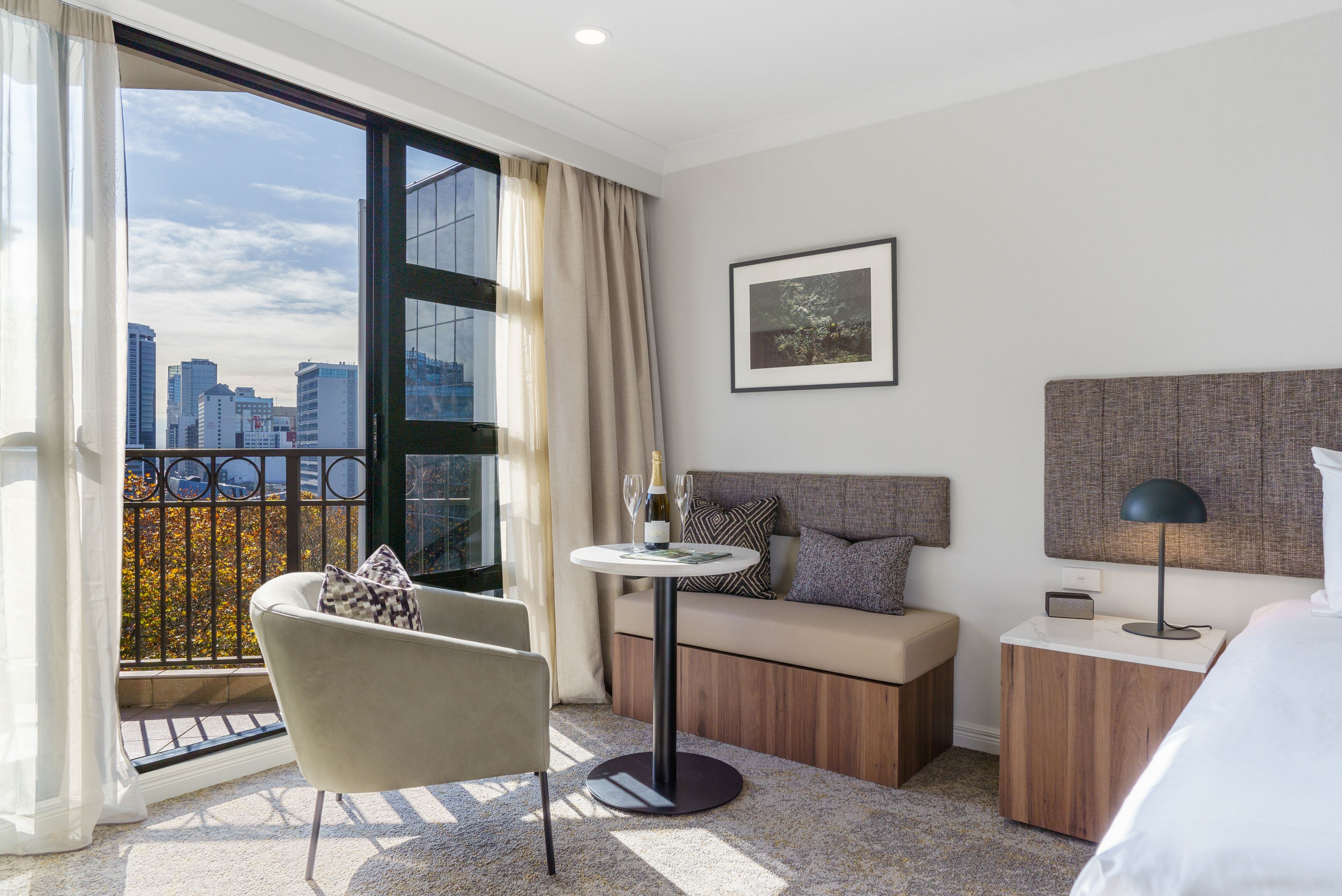 A modern hotel room with a city view, a round table with a bottle and glasses, a sofa with cushions, a bedside lamp, and a framed picture on the wall.