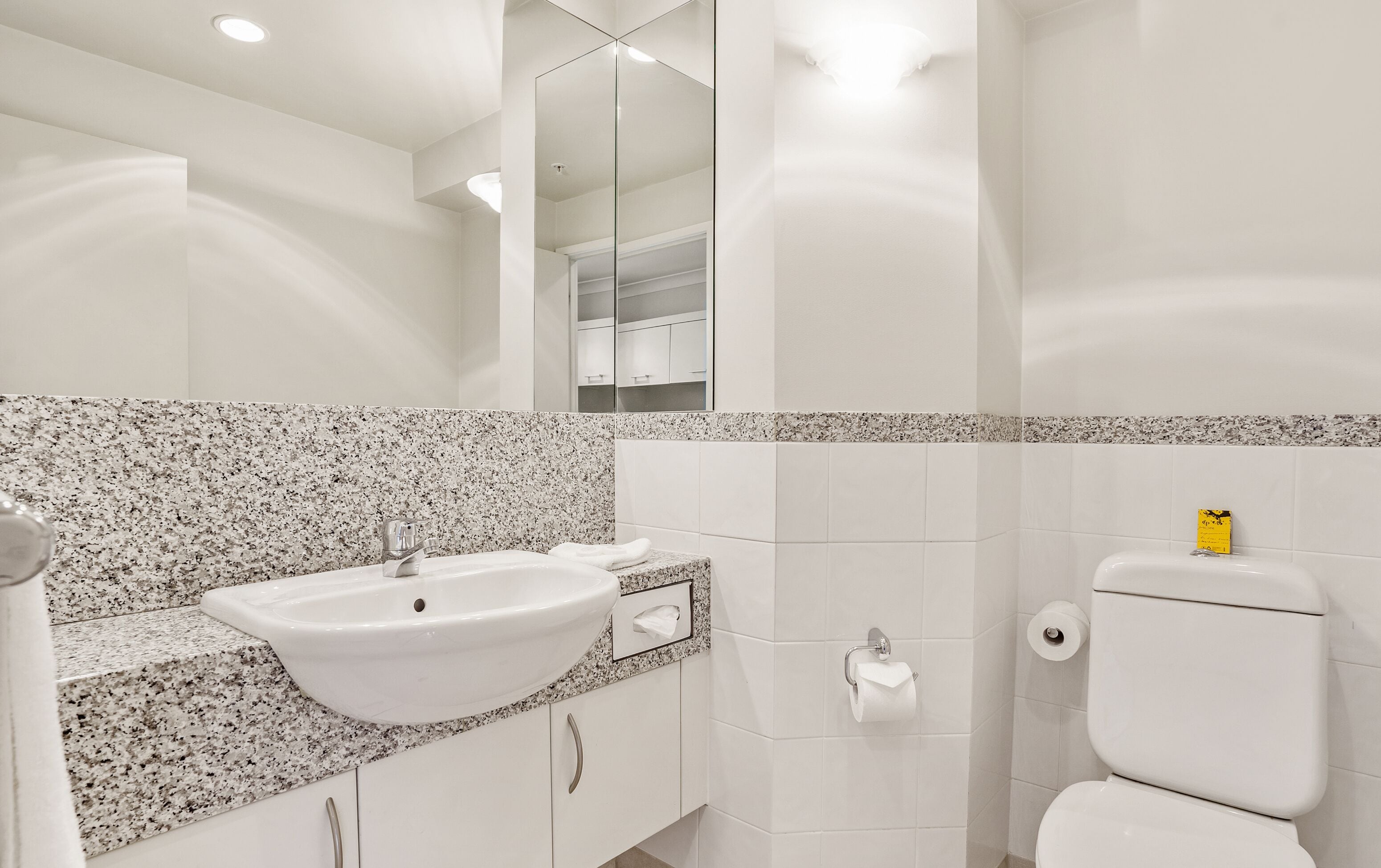 A bathroom with a white sink, granite countertop, white tiles, a toilet, and a mirror.