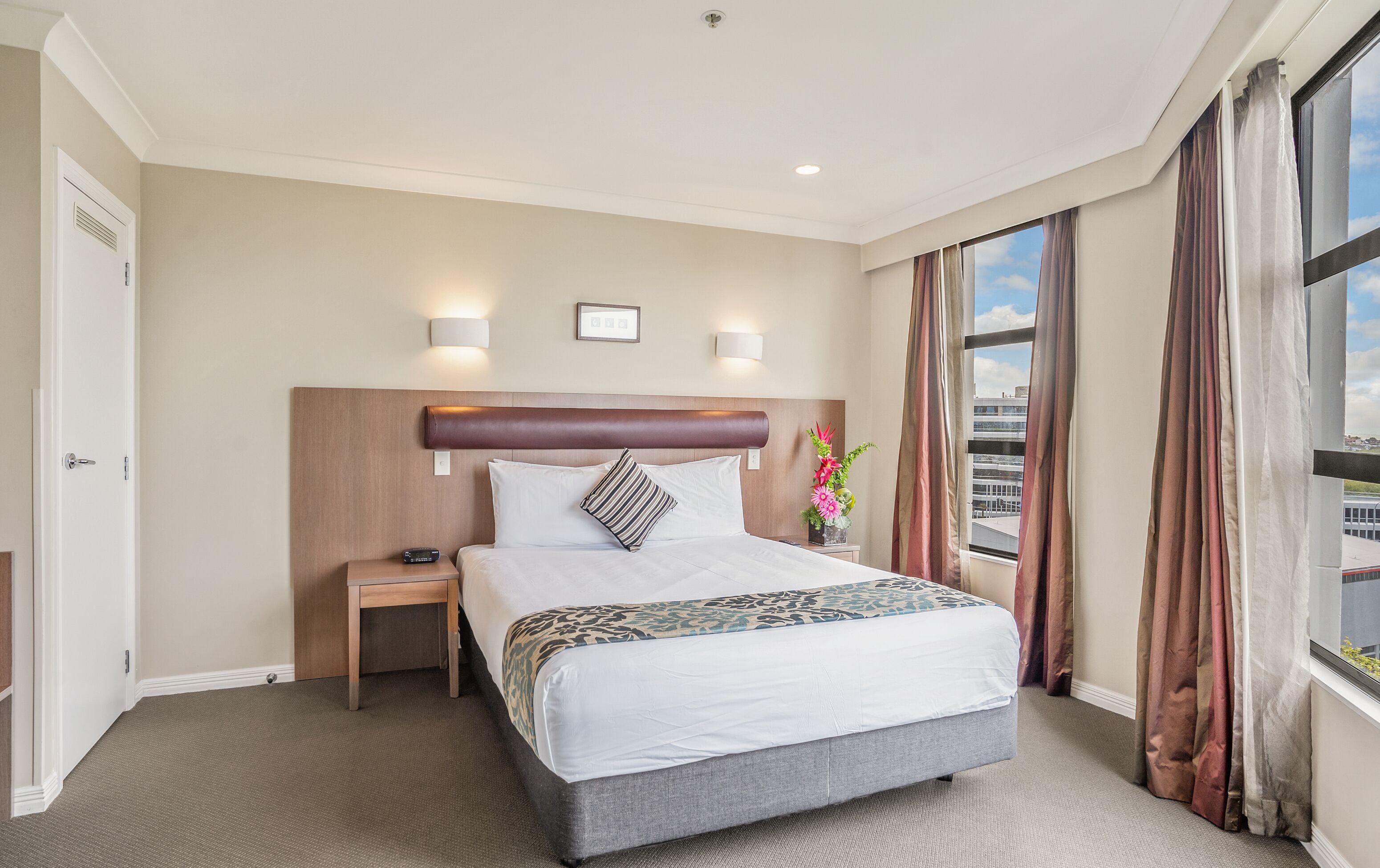 A hotel room with a large bed, a nightstand, two wall-mounted lights, a window with curtains, and a view of the city.