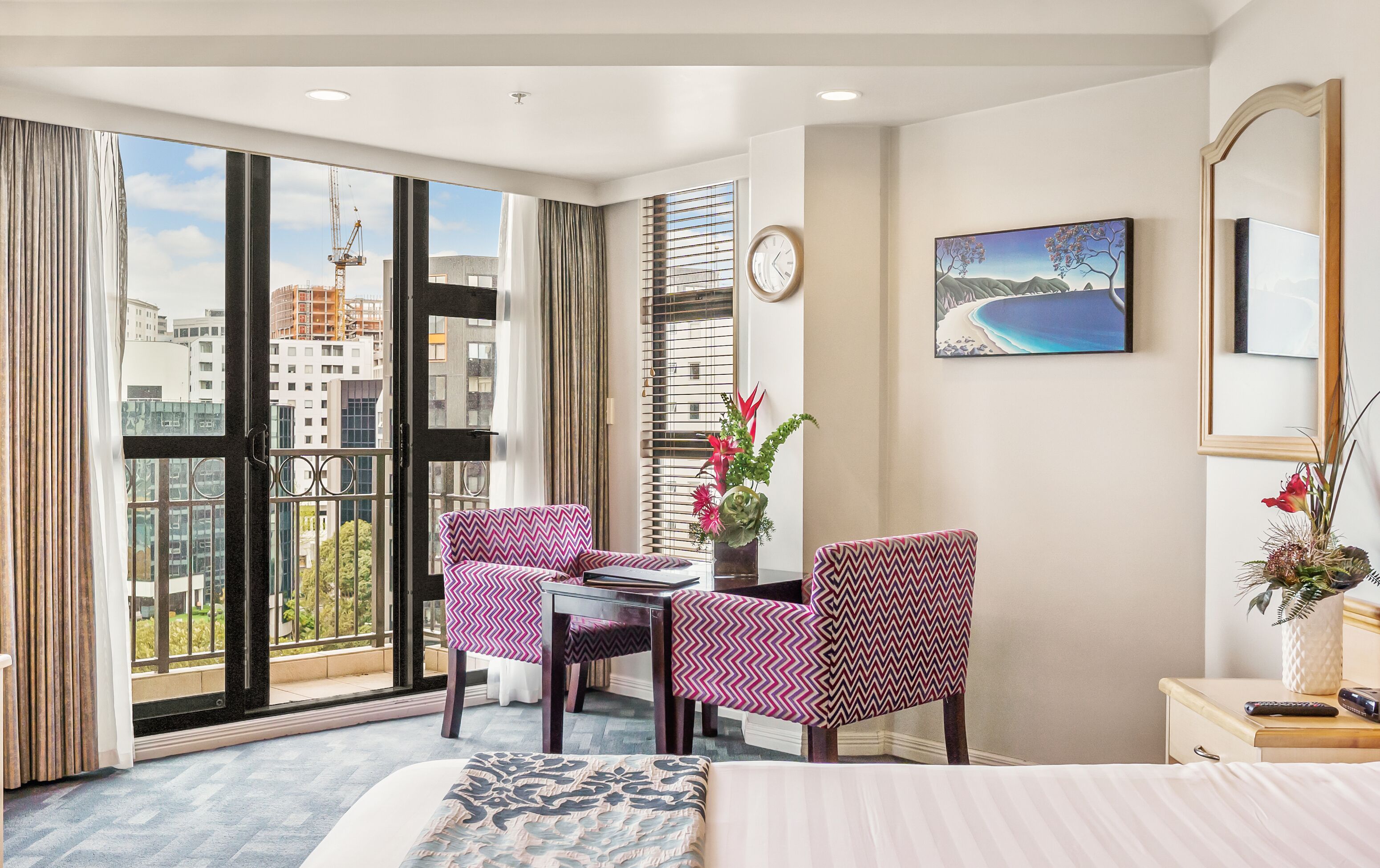 Una habitación de hotel con un ventanal grande, una cama, dos sillones, una mesa, un reloj y un cuadro.