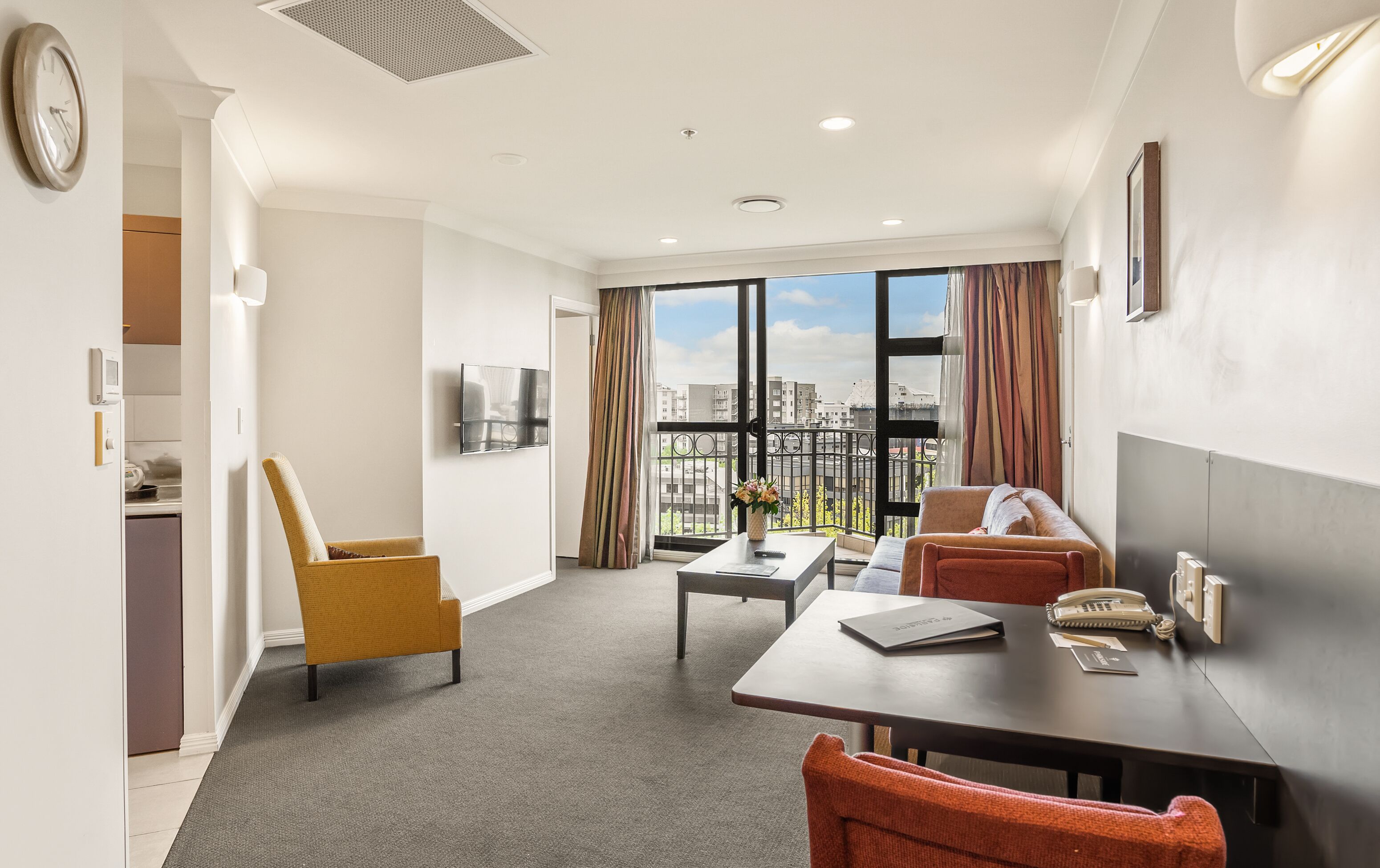 Una habitación de hotel con balcón, zona de comedor, sofá, televisor y kitchenette.