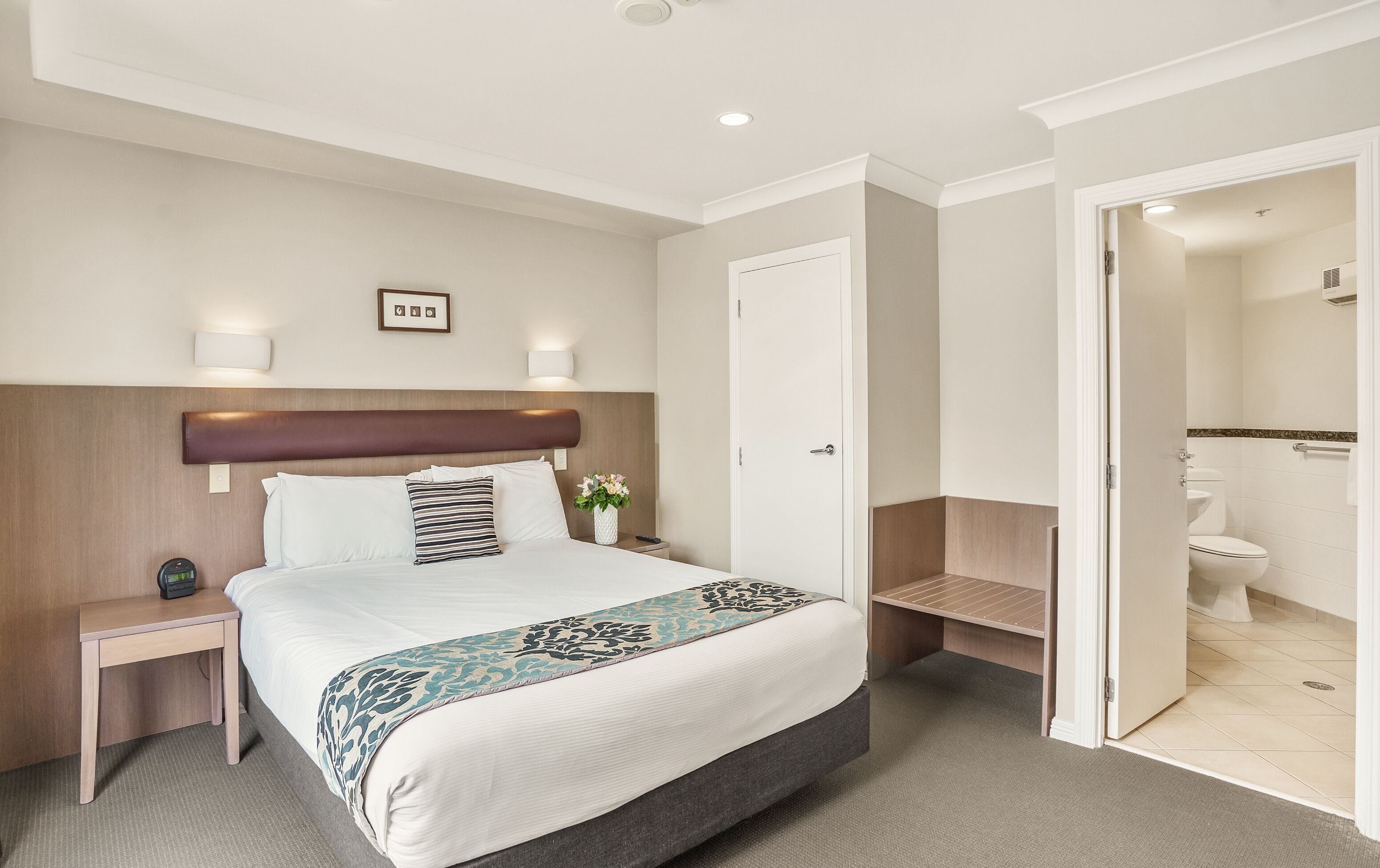 A hotel room with a large bed, bedside tables, a nightstand, a wall-mounted lamp, a framed picture, and a bathroom visible through an open door.