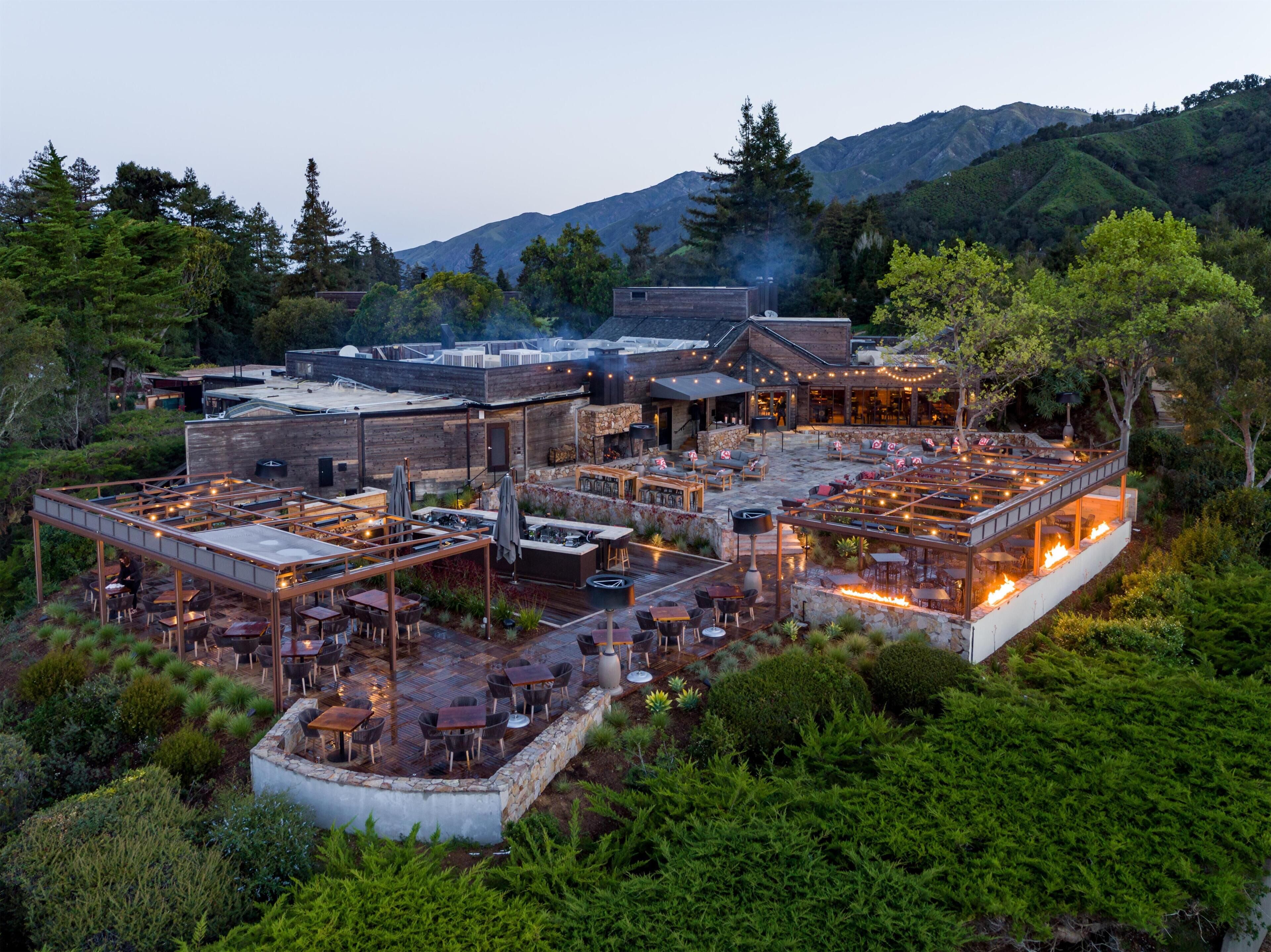 A rooftop restaurant with outdoor seating, a stone fireplace, and a view of the surrounding hills.
