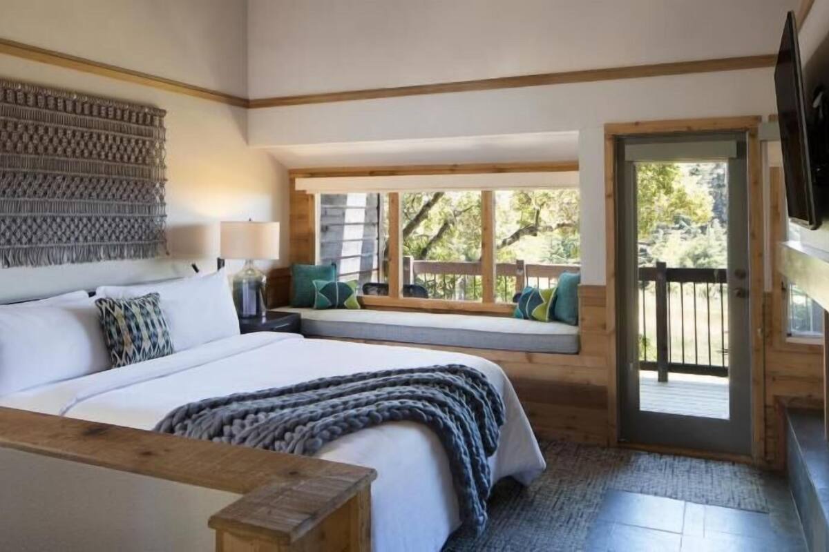 Un dormitorio con una cama grande, cabecera de madera, televisor montado en la pared y un asiento junto a la ventana con vista a árboles.