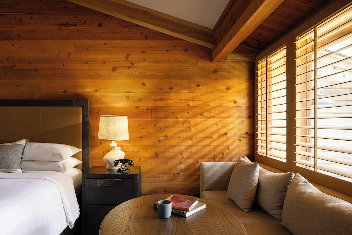 Una habitación acogedora con techo de madera, una cama con ropa blanca, una mesa redonda con un libro y una taza, y una ventana con persianas.