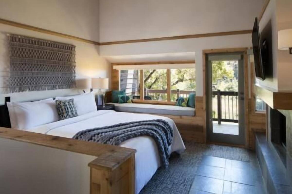 Un dormitorio con una cama grande, un banco de madera, un televisor y un rincón con asiento junto a la ventana que da a una terraza.