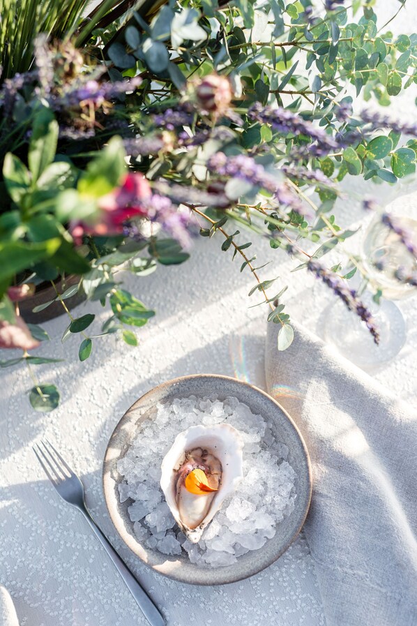 Una mesa de comedor con una fuente de ostras sobre hielo, rodeada de follaje y un mantel blanco.