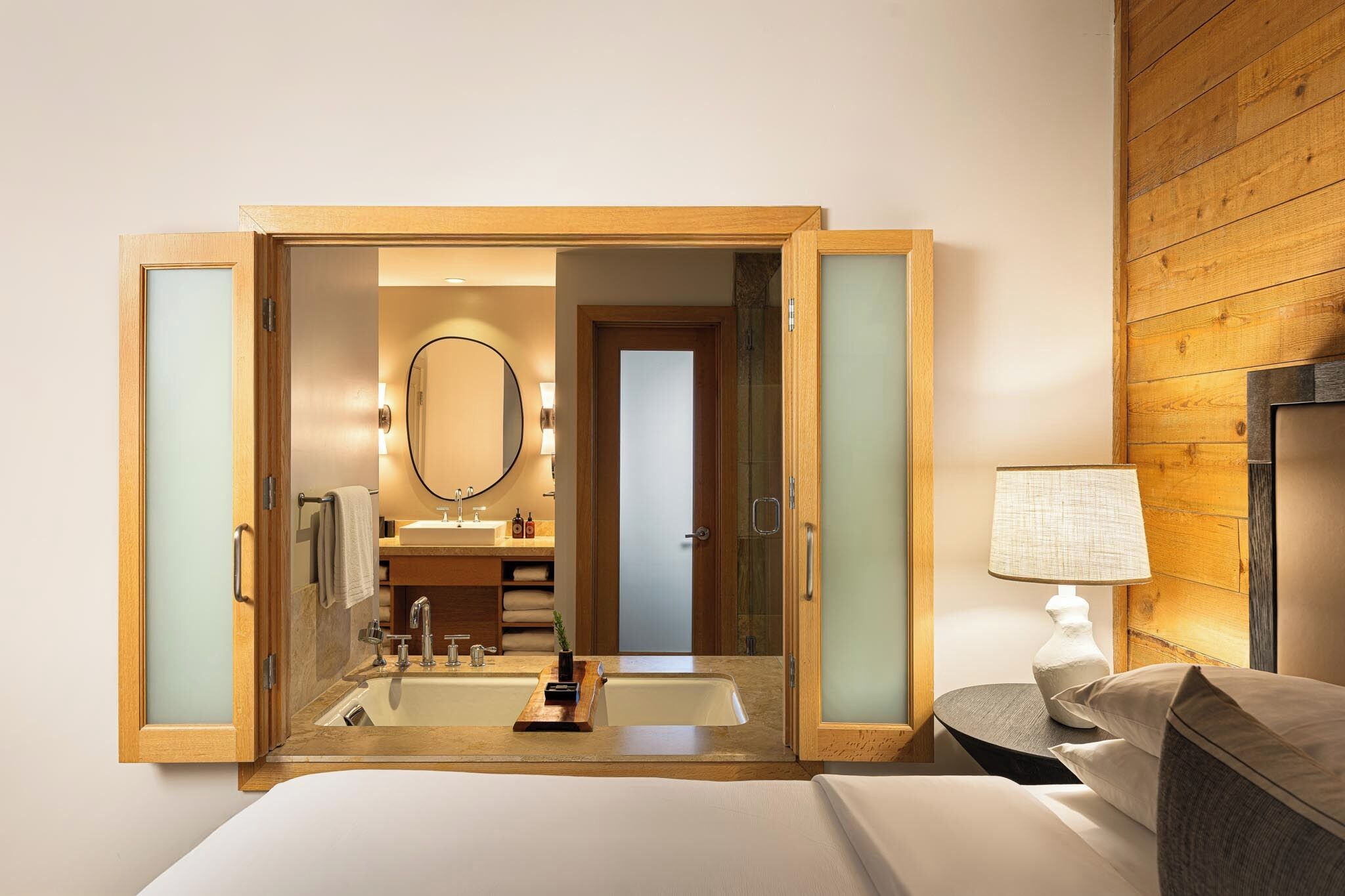 A hotel room with a wooden-framed mirror, a bedside table with a lamp, a bed, and a bathroom visible through an open door.
