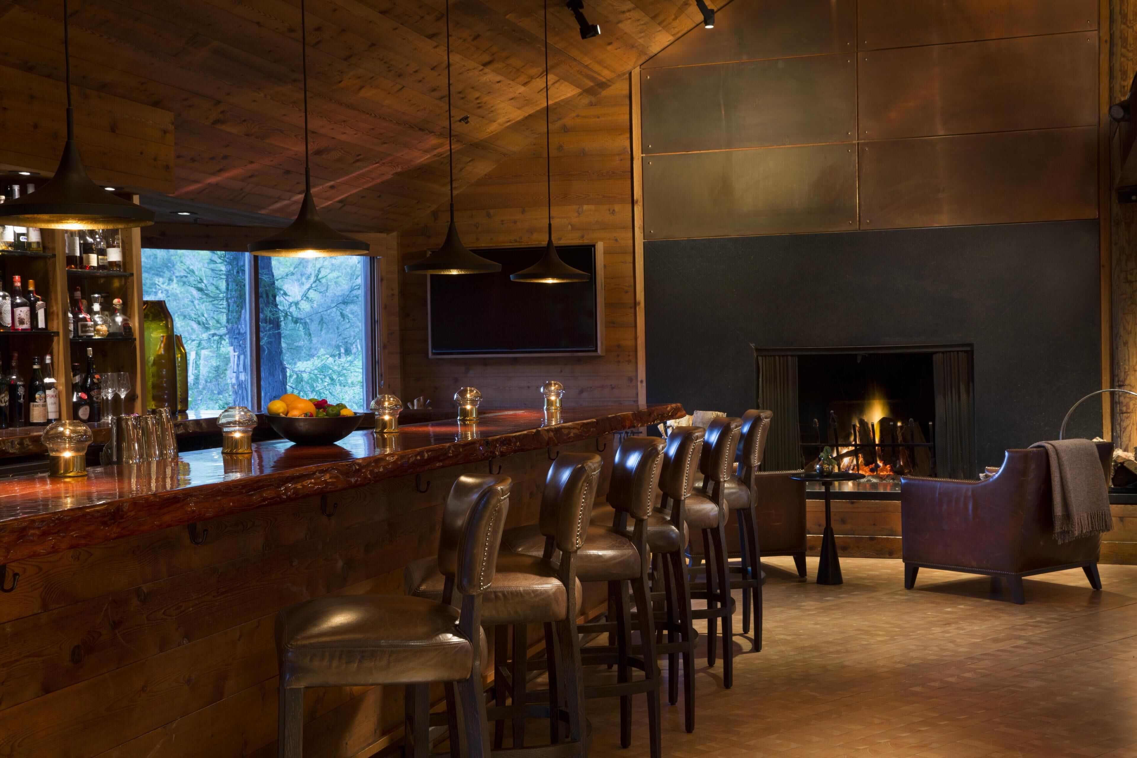 A bar area with wooden ceiling, bar stools, a fireplace, and a view of trees.