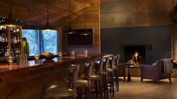 A bar area with wooden ceiling, bar stools, a fireplace, and a view of trees.