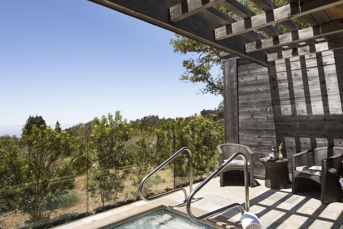 Un jacuzzi con barandas metálicas, una pérgola de madera, mobiliario de mimbre y vistas a árboles y colinas.