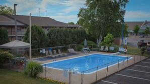 A fenced swimming pool with lounge chairs and a covered area, surrounded by greenery and a parking lot. - Villager Lodge (Niagara Falls)