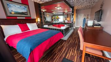 A hotel room with a large bed, a red and blue bedspread, a wooden floor, a red and white striped wall, a large mirror, a red bathtub, a wooden desk, and a chair.