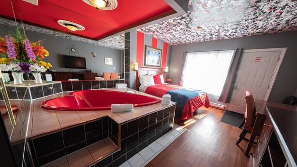 A hotel room with a red and white striped wall, a large window, a red bathtub, a bed with red bedding, and a flat-screen TV on the wall. - Villager Lodge (Niagara Falls)