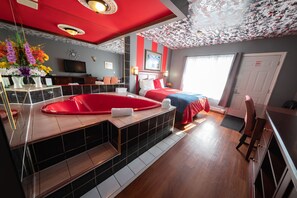 A hotel room with a red and white striped wall, a large window, a red bathtub, a bed with red bedding, and a flat-screen TV on the wall. - Villager Lodge (Niagara Falls)