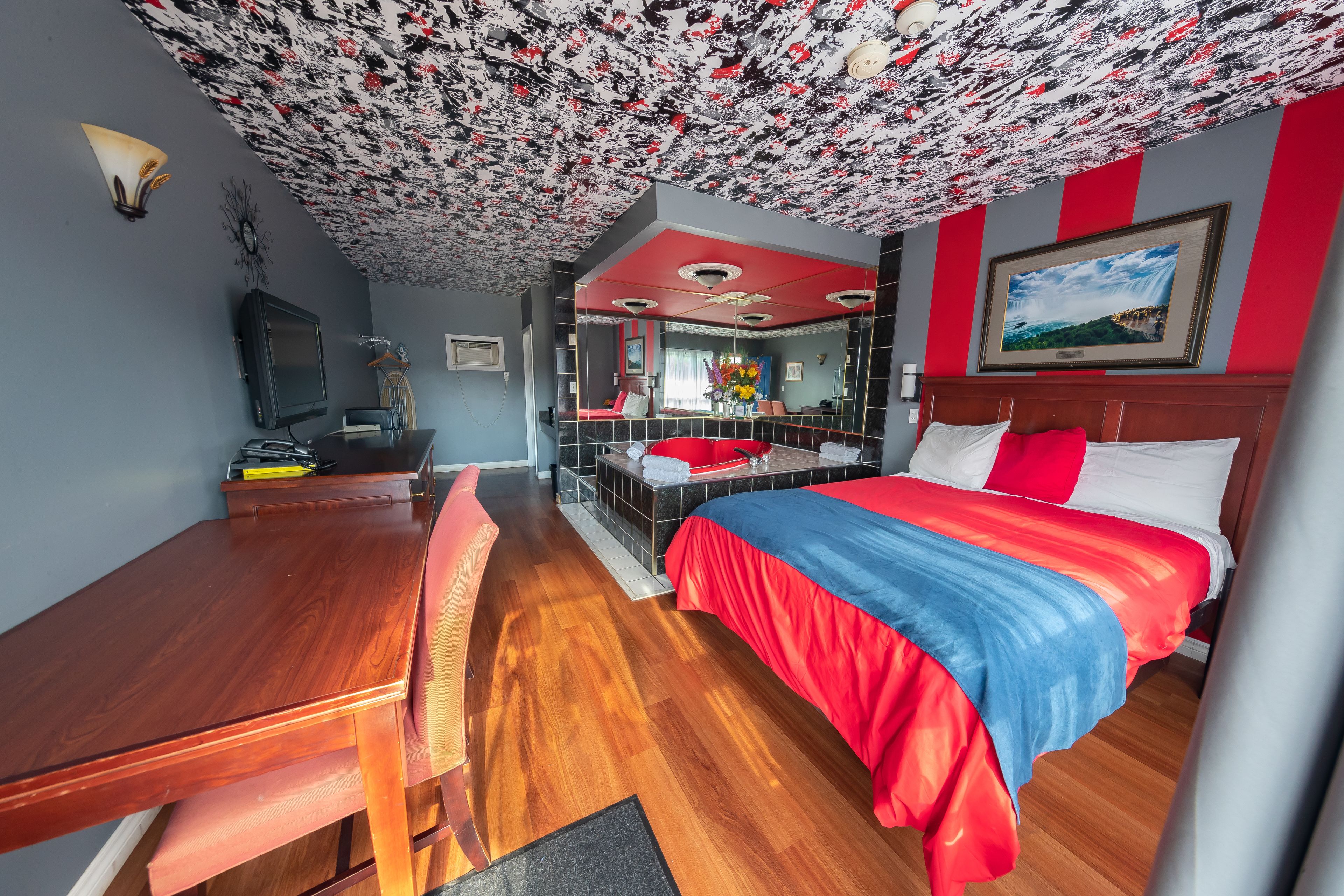 A hotel room with a wooden desk, a bed with red and blue bedding, a ceiling with a floral pattern, and a large mirror.