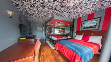 A hotel room with a wooden desk, a bed with red and blue bedding, a ceiling with a floral pattern, and a large mirror.
