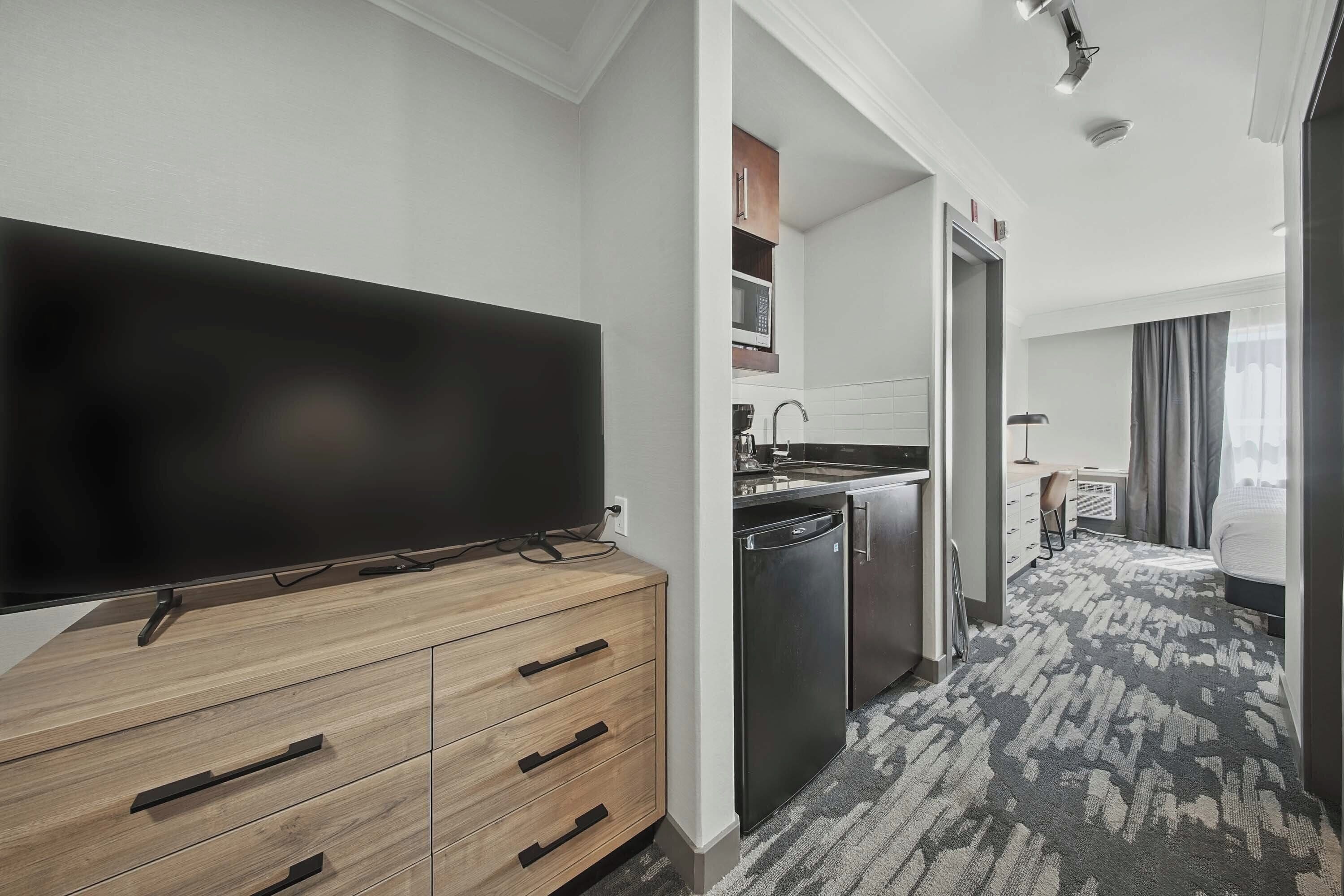 A modern hotel room with a flat-screen TV on a wooden dresser, a kitchen area with a microwave, and a patterned carpet.