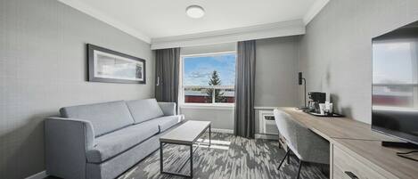 Une chambre d’hôtel moderne avec un canapé gris, une petite table, un bureau avec une chaise, une télévision et une grande fenêtre avec des rideaux.
