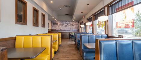A restaurant interior with yellow and blue booths, wooden tables, and a view of the street through large windows.