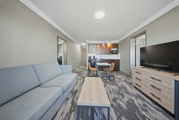 A modern hotel room with a grey sofa, a small wooden table, a TV, and a kitchenette in the background.