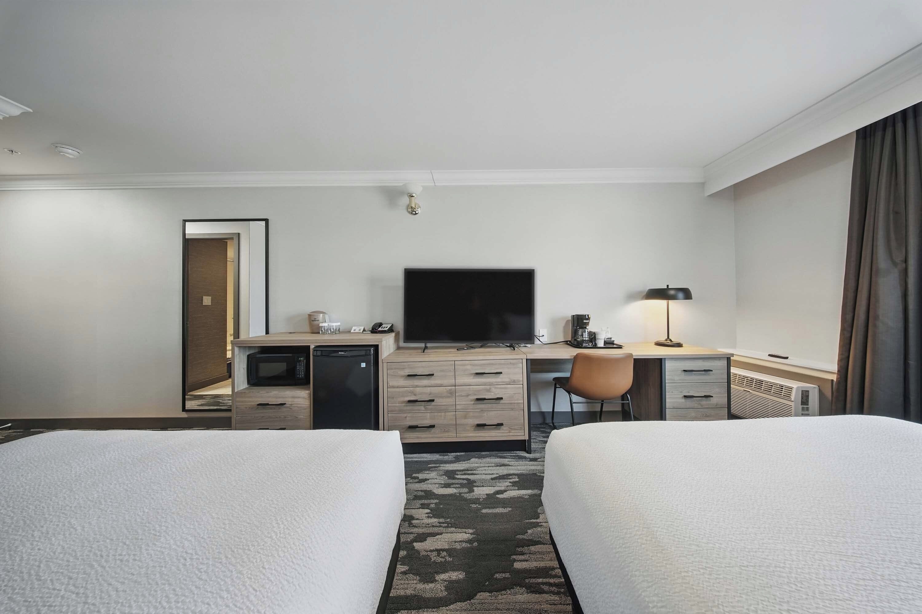 A hotel room with two beds, a desk, a television, a coffee machine, and a small kitchenette.