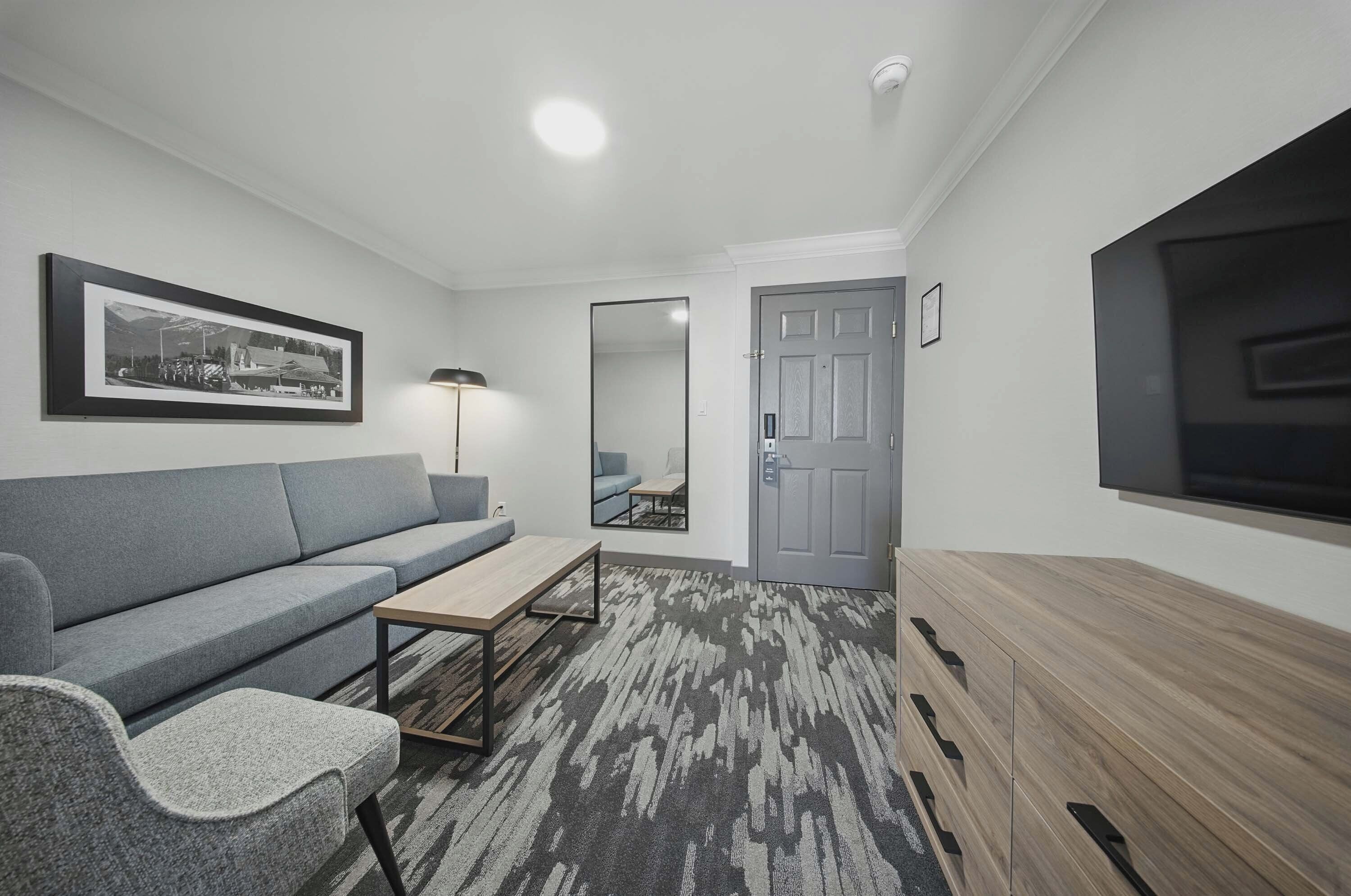 A modern living room with a grey sofa, a wooden coffee table, a patterned rug, and a flat-screen TV mounted on the wall.