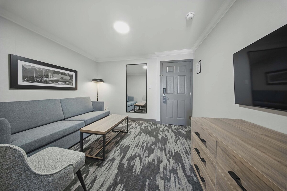 A modern living room with a grey sofa, a wooden coffee table, a patterned rug, and a flat-screen TV mounted on the wall.