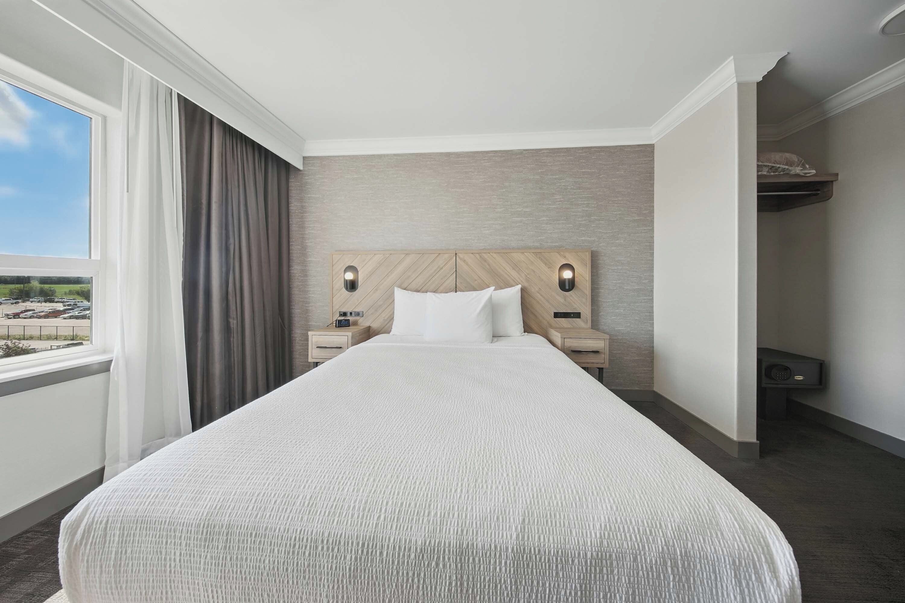 A hotel room with a large bed, two bedside tables with lamps, a view of a parking lot through a window, and a built-in shelf above the bed.