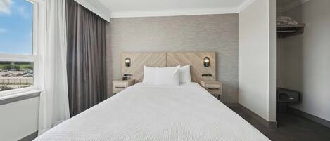 A hotel room with a large bed, two bedside tables with lamps, a view of a parking lot through a window, and a built-in shelf above the bed.