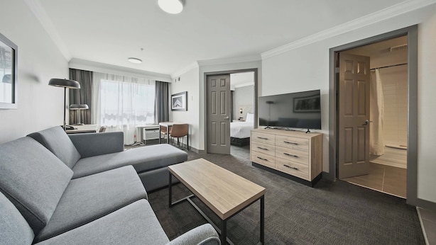 A modern hotel room with a grey sectional sofa, a wooden coffee table, a TV, a desk with a chair, and a wardrobe.