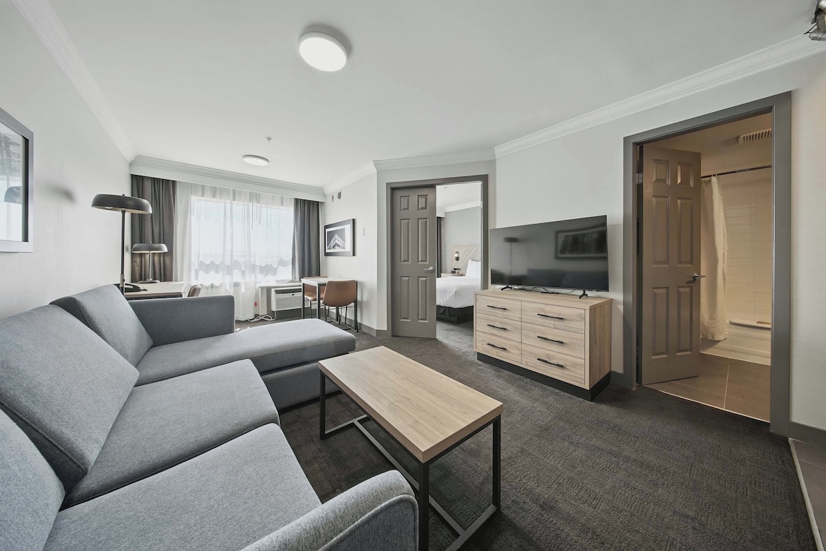 A modern hotel room with a grey sectional sofa, a wooden coffee table, a TV, a desk with a chair, and a wardrobe.