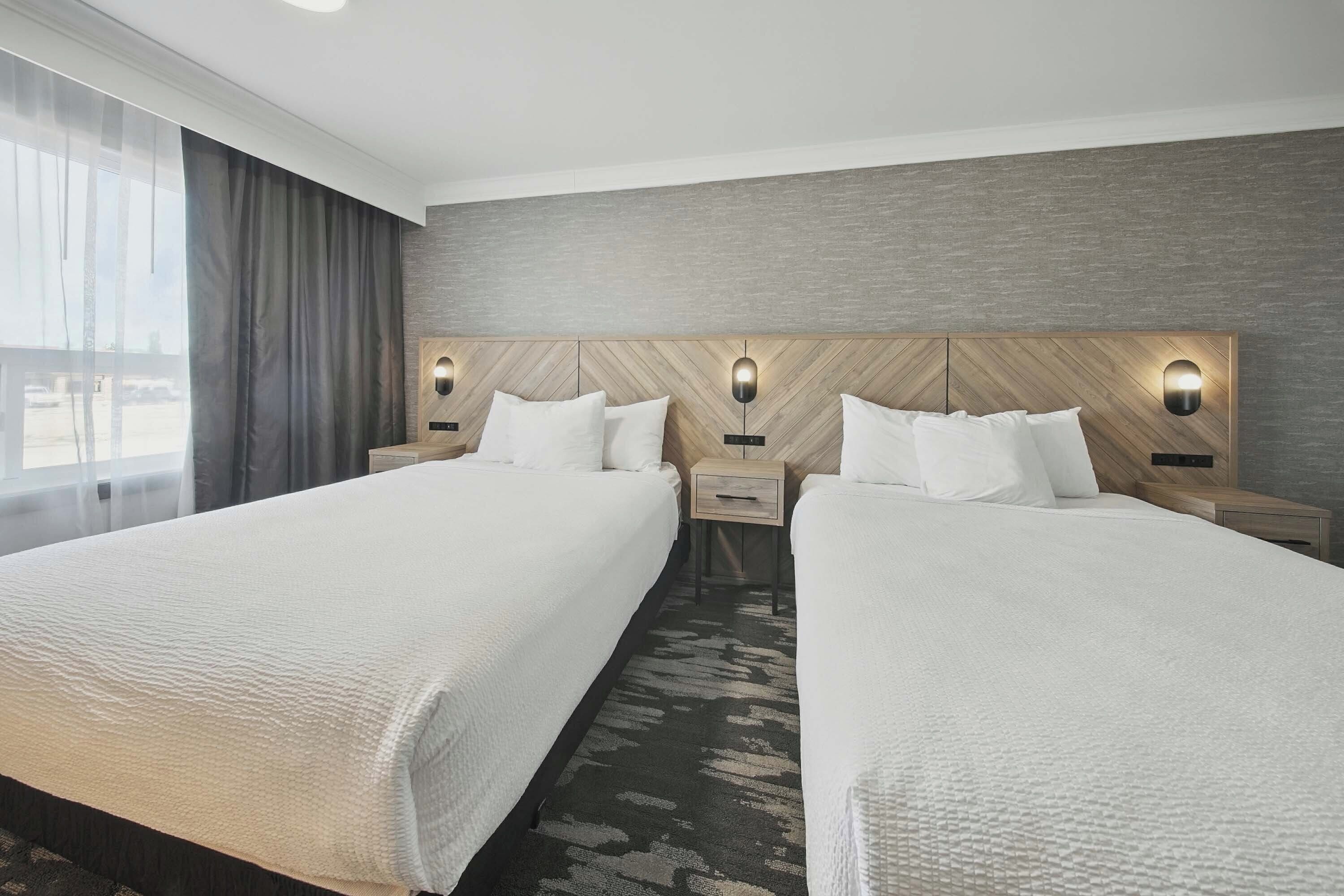 Une chambre d’hôtel avec deux lits, une grande fenêtre avec des rideaux et une tête de lit en bois agrémentée de lampes fixées au mur.