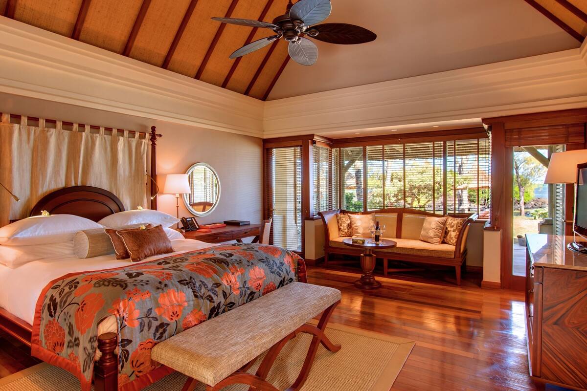 A bedroom with a large bed, a wooden ceiling fan, a bedside table with a lamp, a chair, a sofa, and a television.
