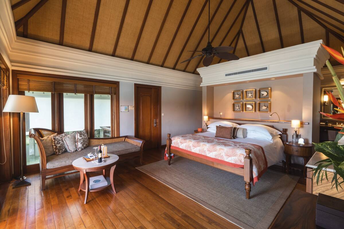 A spacious bedroom with a large bed, a sitting area with a sofa and a small table, wooden flooring, and a ceiling fan.
