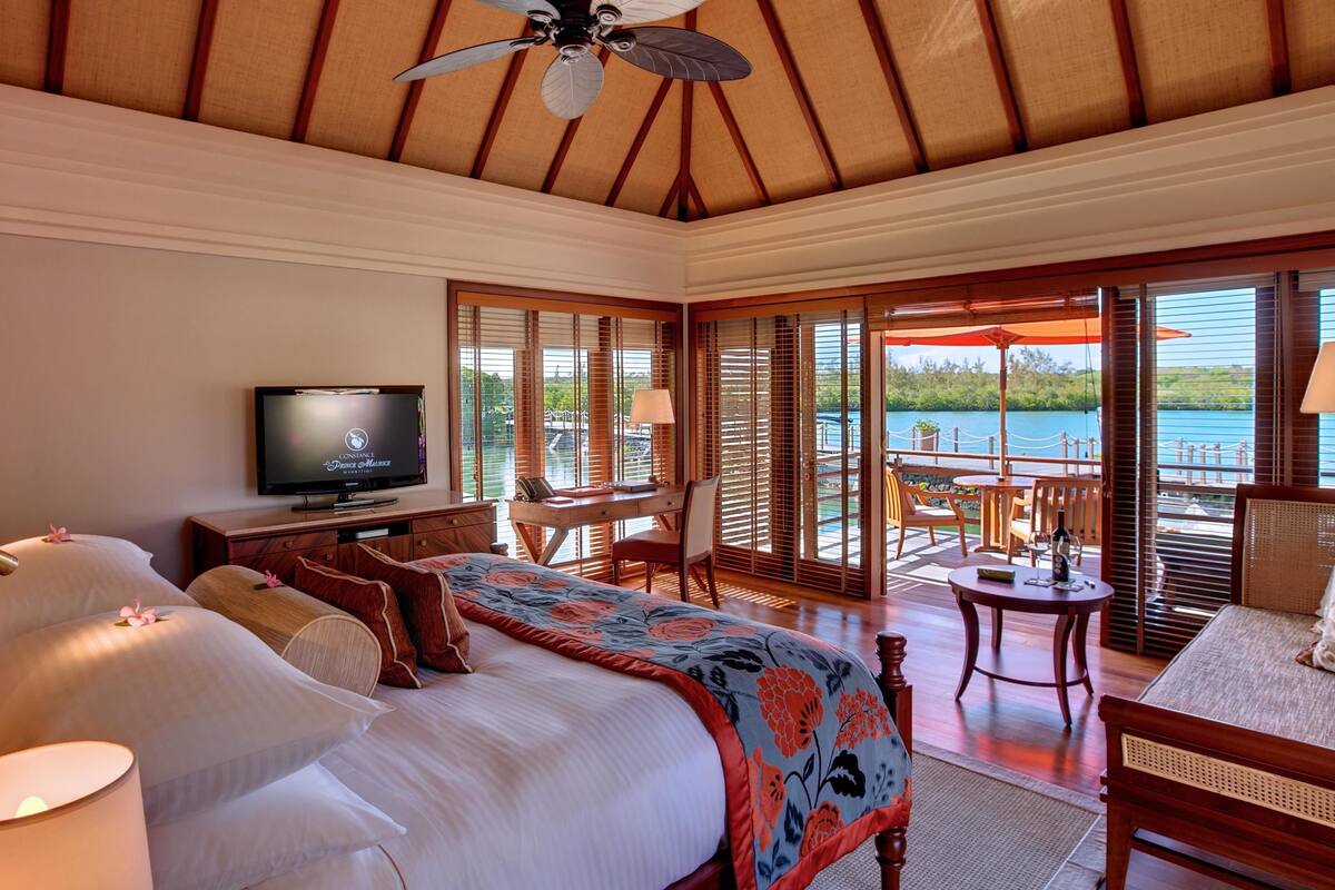 A bedroom with a bed, a desk, a television, and a balcony with a view of the water.