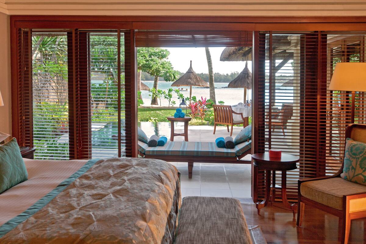 A hotel room with a bed, a small table, a chair, and a view of the beach and palm trees.