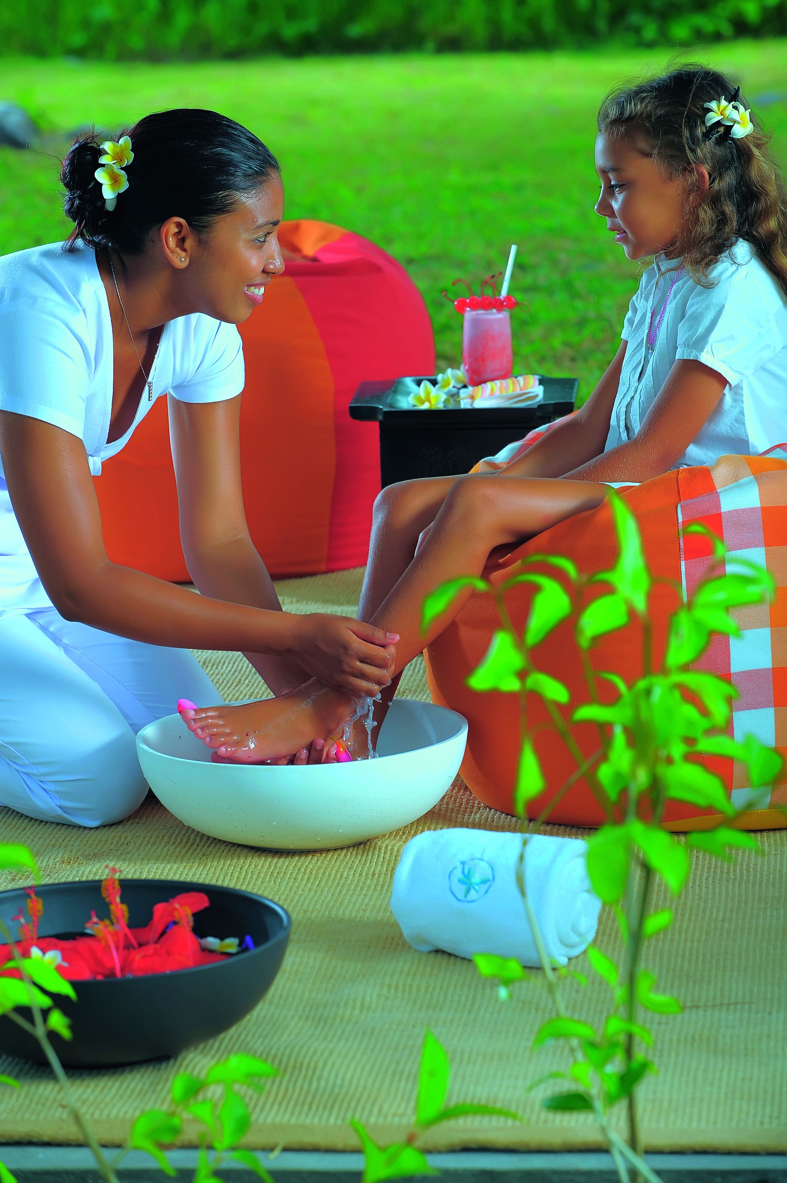 A person washing another person's feet in a bowl, with a plant and a red cushion in the foreground.