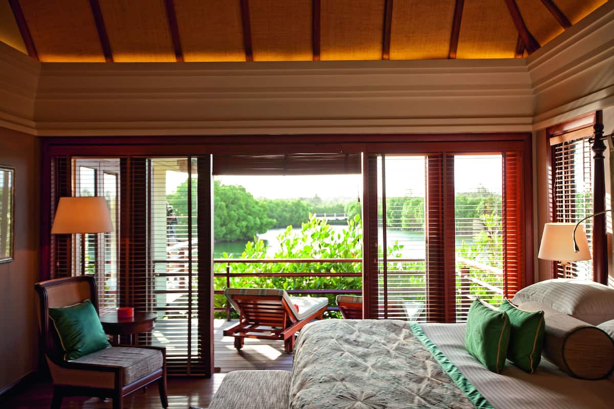 A hotel room with a bed, a chair, a table, and a view of greenery and water outside.
