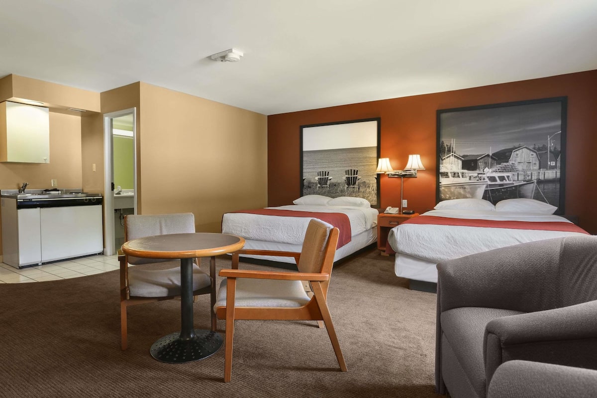 A hotel room with two beds, a dining table, a chair, a small kitchen area, and a large framed picture on the wall.