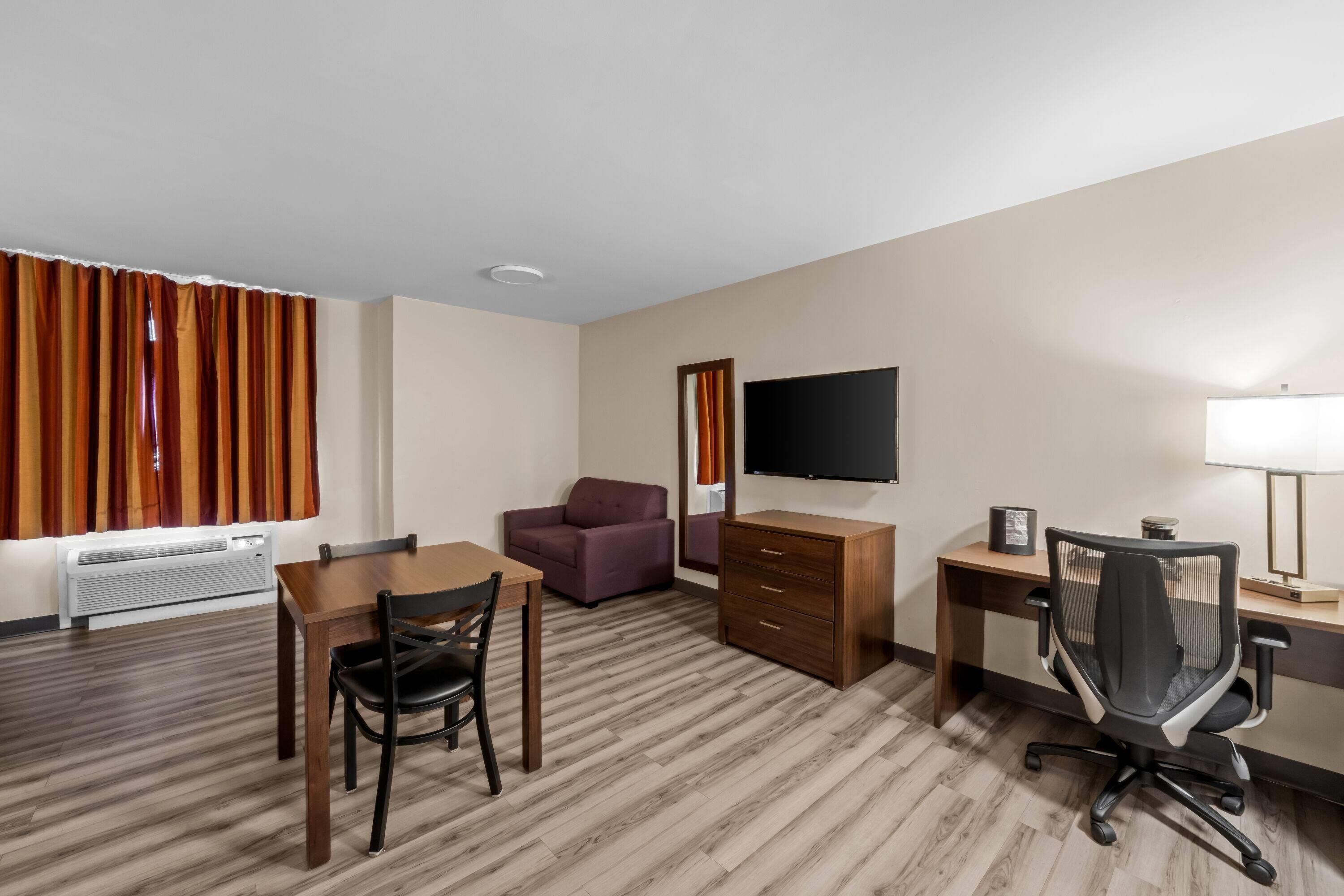 Una habitación de hotel con piso de madera, una mesa de comedor con sillas, un sofá, un televisor, un escritorio con una silla y una lámpara.