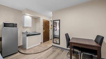 Cocina compacta con microondas, lavabo y armario, mesa de comedor con sillas y un espejo que refleja un dormitorio.
